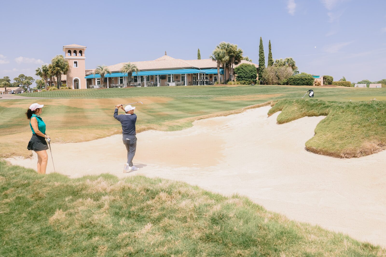 Billy Belair hitting from a greenside bunker with a student watching, Grande Dunes clubhouse in the background